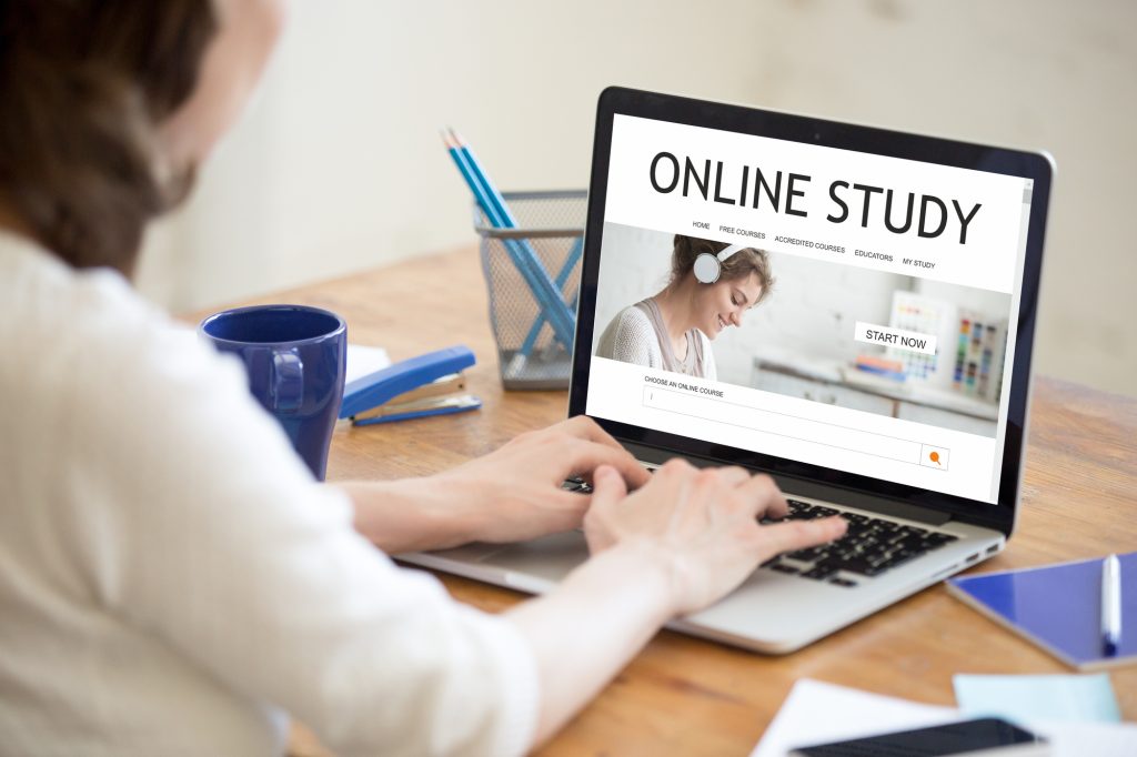 Young happy woman working in office interior, browsing e-learning website. Attractive female studying online using web service on laptop. Back view over the shoulder. On-line education concept